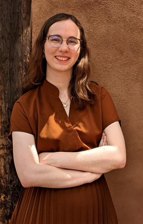 Merritt Zamboni wearing a brown dress and glasses, crossing her arms and smiling.